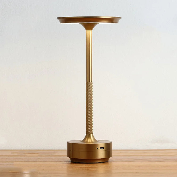 A gold cordless lamp standing on a pale wooden desk